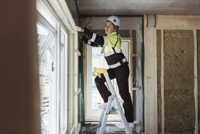 Full length of female building contractor measuring window frame standing on step ladder at construction site