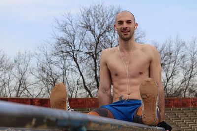 Portrait of shirtless man listening music while exercising against sky