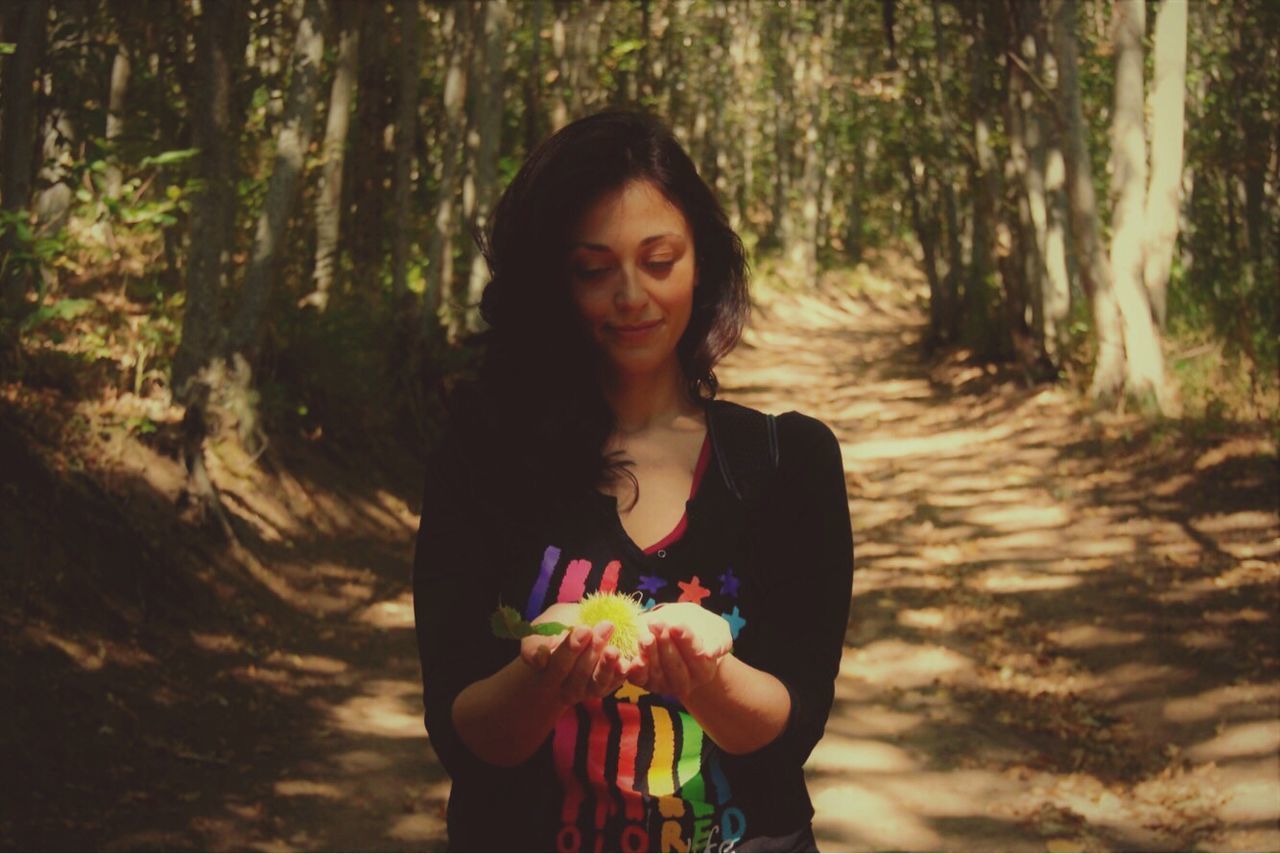 SMILING YOUNG WOMAN STANDING BY TREE IN FOREST