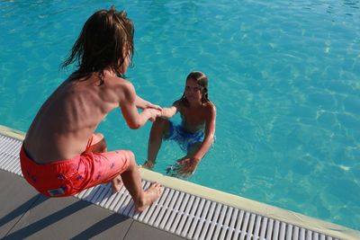Rear view of shirtless boy pulling friend from swimming pool