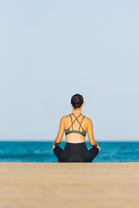 Rear view of woman meditating at beach against clear sky