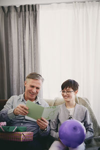 Smiling grandfather reading greeting card while sitting with grandson and gifts at party in living room