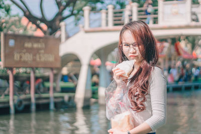 Portrait of young woman eating food while standing against river and text on board
