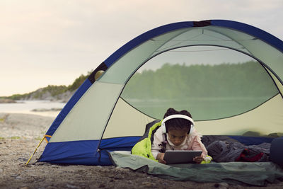 Girl with headphones using digital tablet while lying in tent at beach against sky during sunset