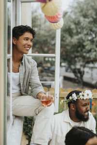 Smiling young man holding wineglass while sitting on window sill during dinner party at cafe