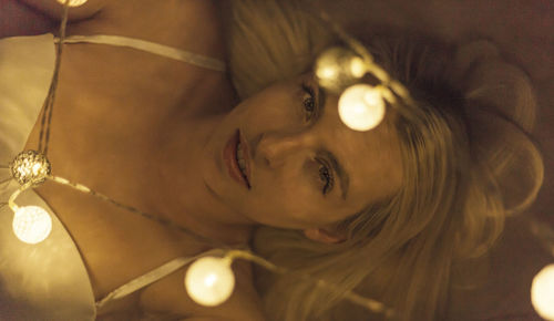 High angle portrait of woman lying on bed seen through illuminated string lights