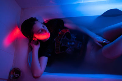 Depressed woman thinking about suicide, lying in clothes in bathtub under neon light