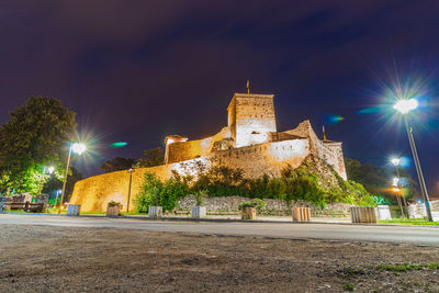 Illuminated street amidst buildings against sky at night