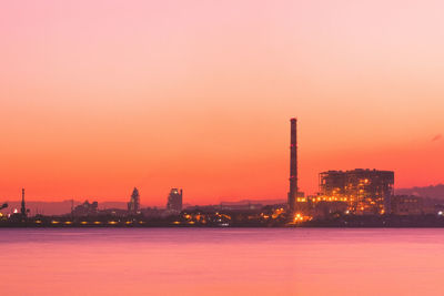 Illuminated factory by river against sky during sunset