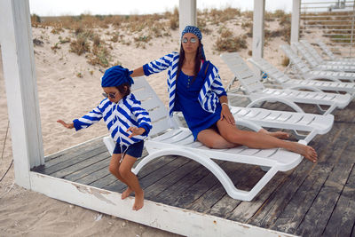 Mother and her baby son in striped blue jackets walk along the beach next to a wooden gazebo