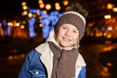 Happy little boy in warm jacket and knitted hat on street in the evening