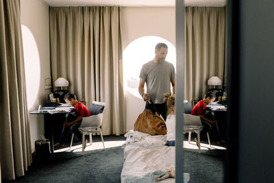 Man holding bag by bed while boy sitting at table in hotel