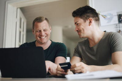 Smiling homosexual couple paying bill through laptop on dining table at home