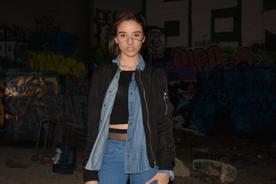 Portrait of young woman standing against graffiti wall at night