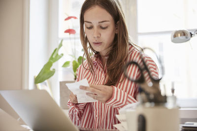 Young woman examining financial bill while using laptop at home
