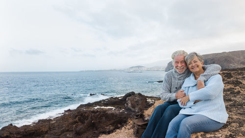 Portrait of smiling couple sitting by sea against cloudy sky