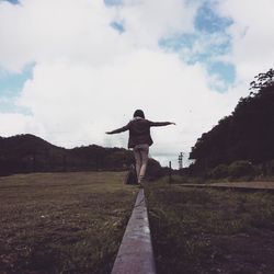 Rear view of woman with arms outstretched walking on railroad track against cloudy sky