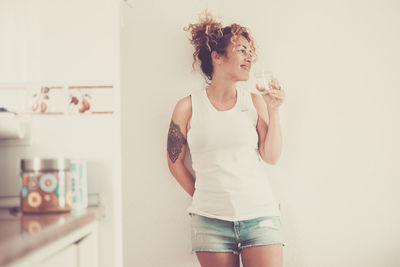 Smiling woman drinking water standing against wall at home