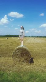 Full length of young woman standing on hay bale at field against sky