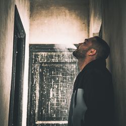 Side view of thoughtful man emitting smoke leaning on wall in old house