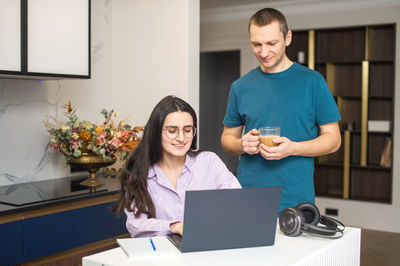 A girl with glasses is sitting and looking at a laptop, a man is standing next to her