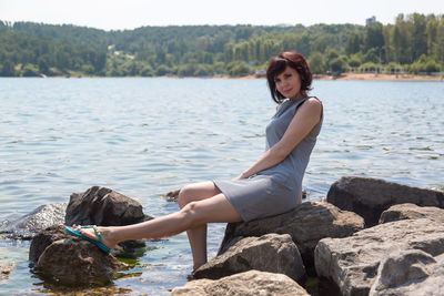 A happy adult woman is sitting rock on the riverbank sunbathing on a sunny day.