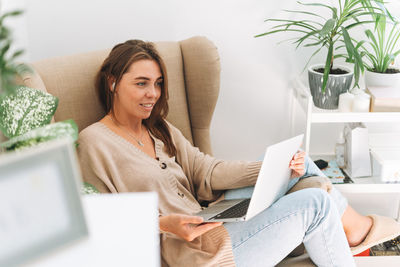 Young smiling attractive woman in cozy beige cardigan working at laptop at home with green house