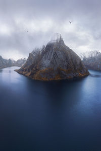 Aerial view of calm water of sea washing island with rocky formations covered in snow under cloudy sky