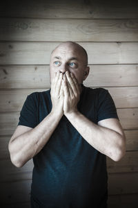 Man with hands covering mouth standing against wooden wall