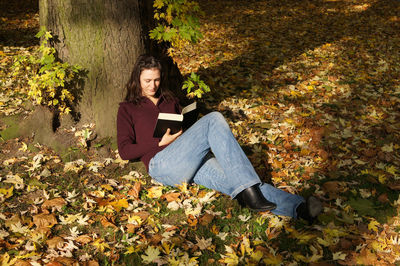 Young woman reading book while leaning on tree during autumn at herrenhausen gardens