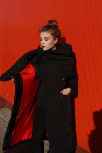 Fashionable teenage girl wearing black warm clothing standing against red wall during sunny day