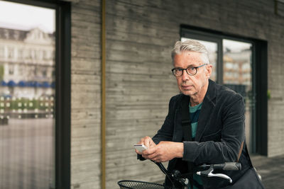 Portrait of senior man using smart phone while standing with bicycle in city