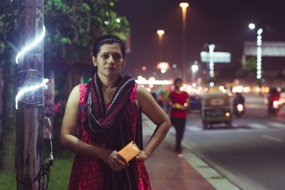 Portrait of young woman standing on illuminated street at night