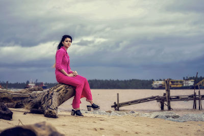 Full length of teenage girl sitting on driftwood at beach against cloudy sky