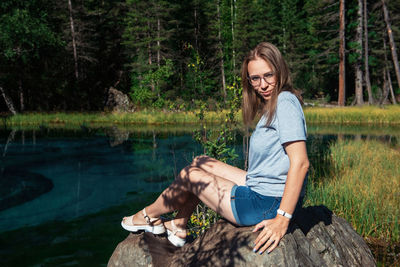Beautiful young woman sitting by lake in forest