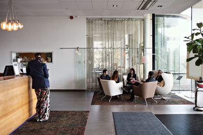 Businesswoman standing at reception with colleagues discussing in while sitting on armchairs in office