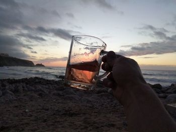 Hand holding glass of water at beach against sky during sunset