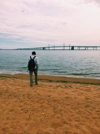 Rear view of man with backpack walking at beach against cloudy sky