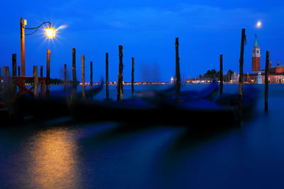 Boats moored on grand canal by illuminated street light against blue sky at night