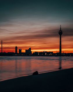 Silhouette buildings by river against sky during sunset in city