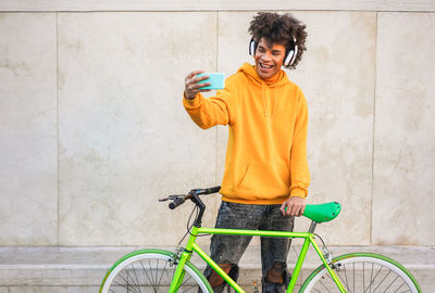 Young man with bicycle taking selfie while standing against wall