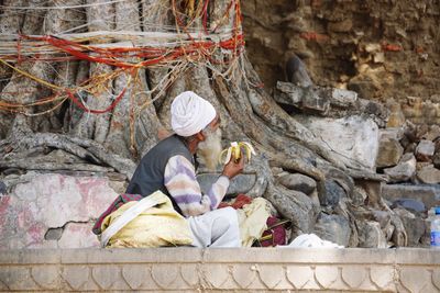 Side view of man eating banana while sitting against tree trunk