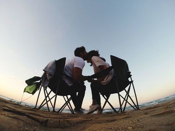 Low angle view of man kissing woman while sitting on chair at beach against clear sky