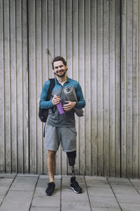 Full length portrait of man with disability carrying exercise mat and water bottle in front of wall