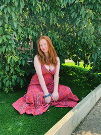 Portrait of young woman sitting on grass against trees at park