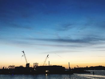 Silhouette cranes at harbor against blue sky during sunset