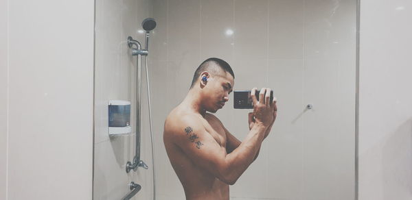 Reflection of shirtless man photographing with mobile phone on mirror in bathroom