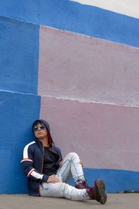 Man sitted on the floor wearing a  hoodie and sunglasses with a colorful wall as background.