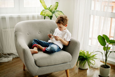 A cute boy is sitting in an armchair and playing a game on his phone or watching educational videos.