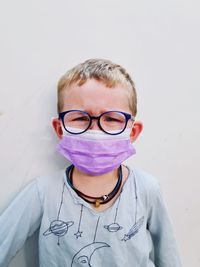 Portrait of boy wearing eyeglasses standing against wall with face mask